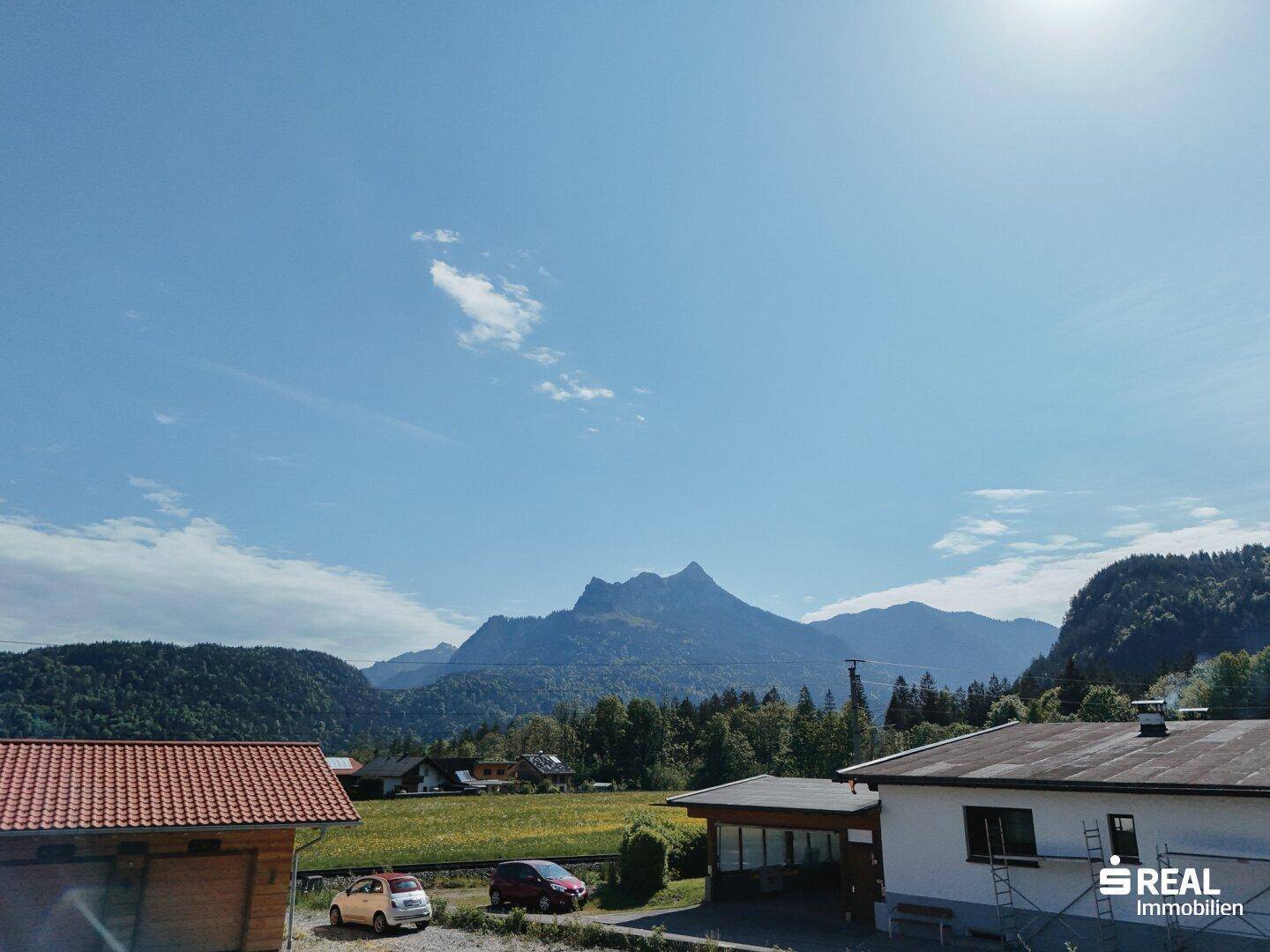 Panoramablick auf eine idyllische Berglandschaft mit weiten Wiesen und Häusern unter strahlendem Sonnenschein.
