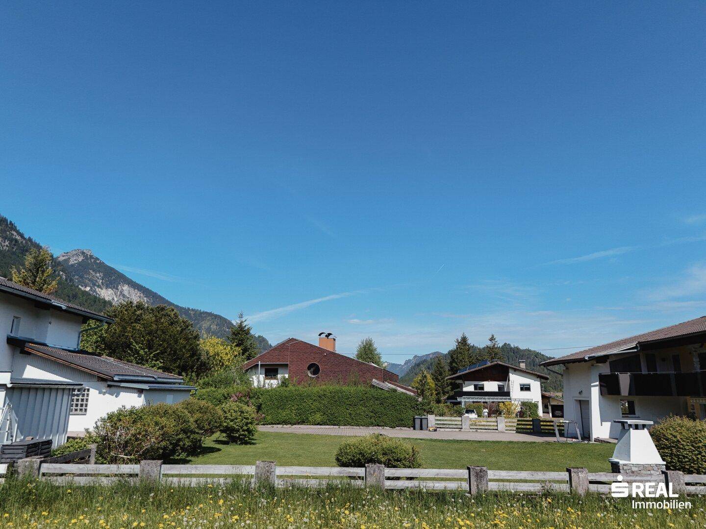 Ansicht von Wohnhäusern mit gepflegten Gärten und einer Bergkulisse im Hintergrund an einem sonnigen Tag.