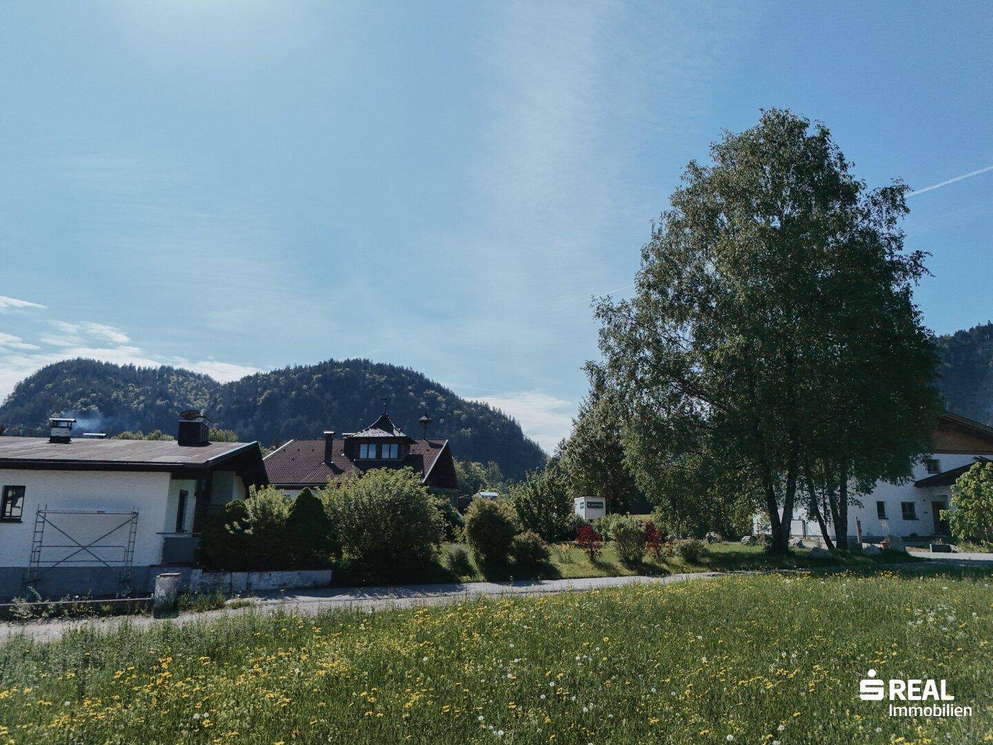 Ländliche Szenerie mit grünen Feldern, Bäumen und Wohnhäusern vor einer sanften Hügellandschaft.