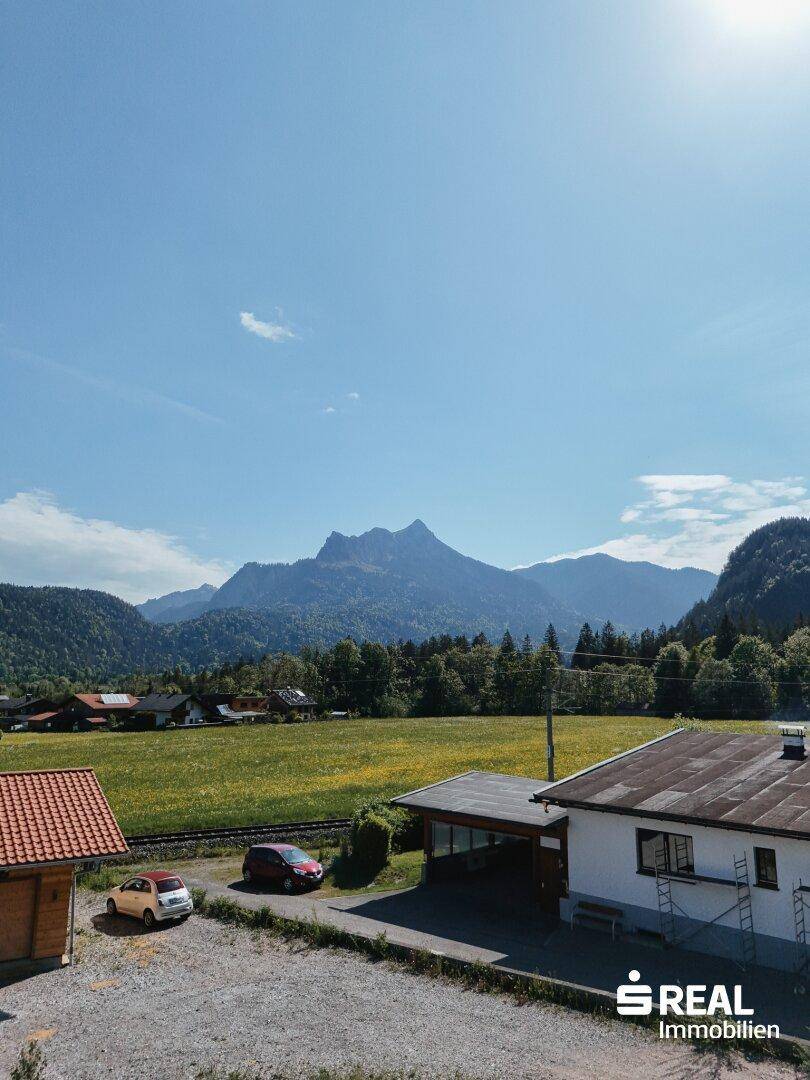 Landschaftsbild mit majestätischen Bergen, grünen Wiesen und einigen Häusern unter blauem Himmel.
