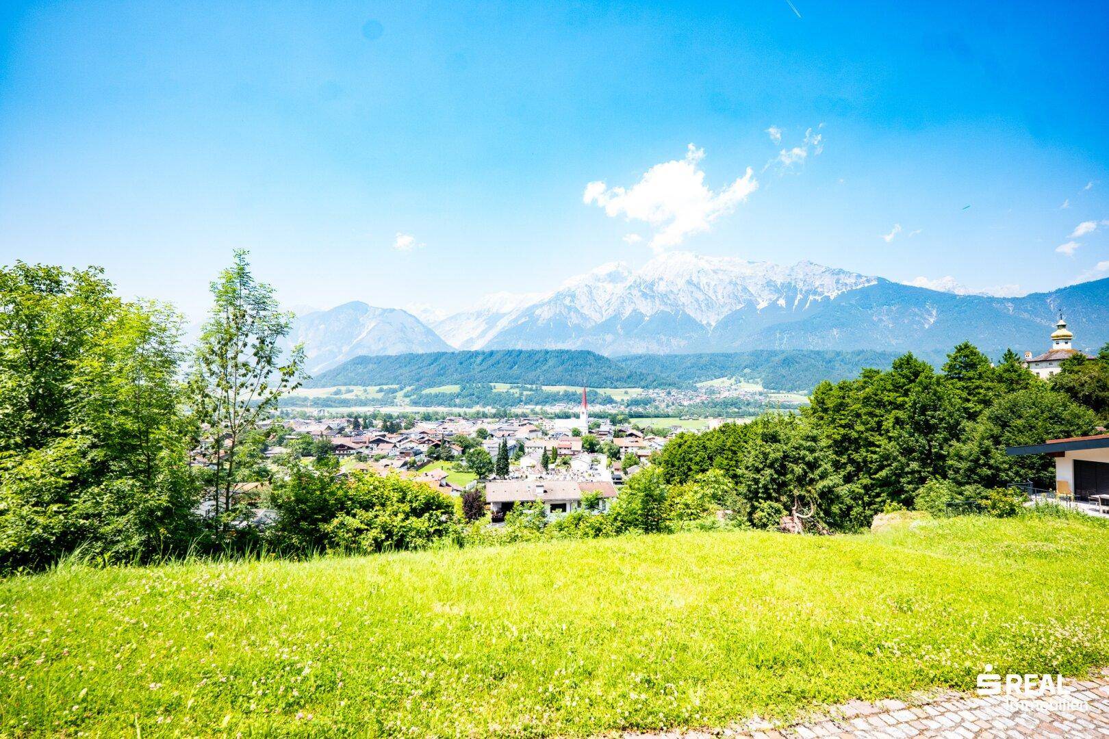 Sonniger Ausblick von einer grünen Wiese auf eine Stadt und die beeindruckende Berglandschaft dahinter.