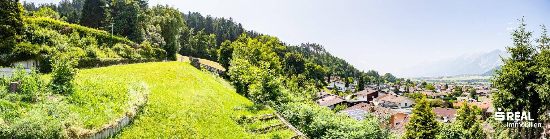 Weitläufiger Panoramablick über grüne Hügel, bewaldete Hänge und eine Siedlung mit Bergkulisse.