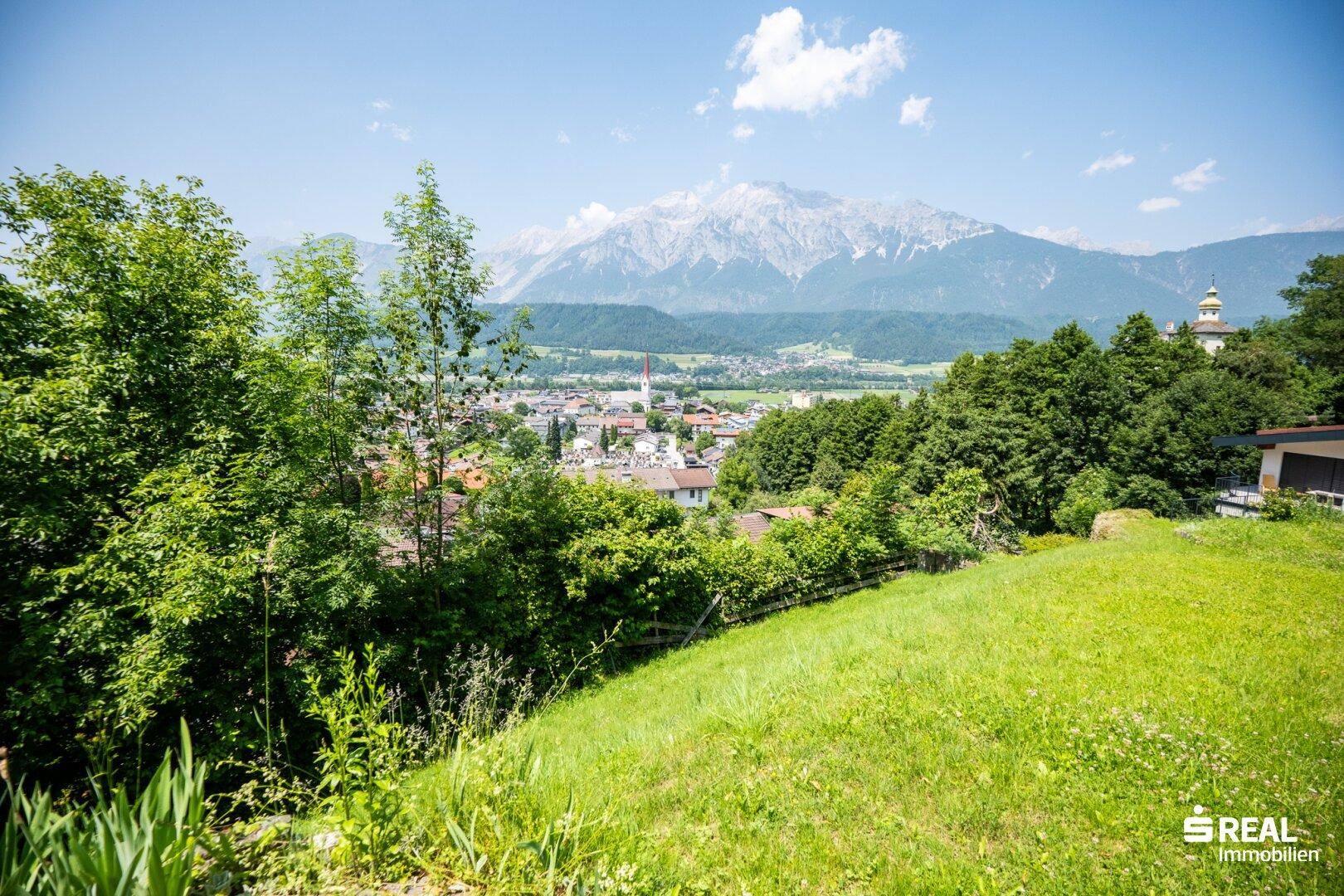 Aussicht von einer grünen Anhöhe mit Bäumen auf eine Siedlung und die majestätische Bergkulisse.