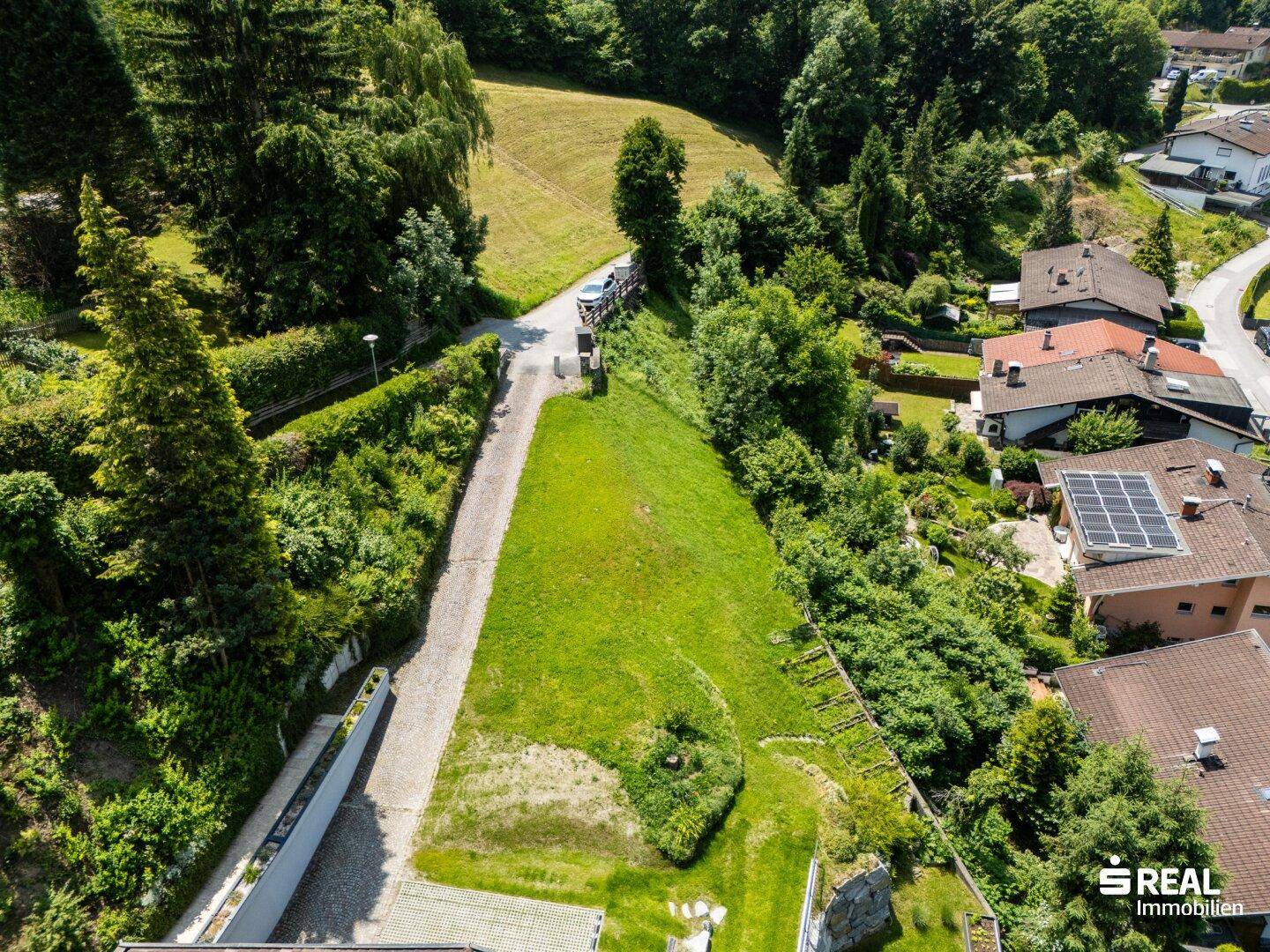 Luftaufnahme eines Grundstücks mit Zufahrt, umgeben von grünen Hügeln und benachbarten Häusern.