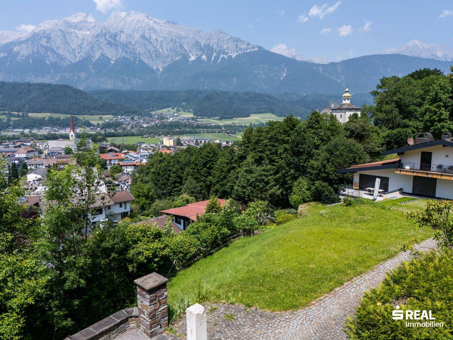 Blick von einer Anhöhe auf ein Haus, grüne Gärten, eine Stadt und die beeindruckende Berglandschaft.