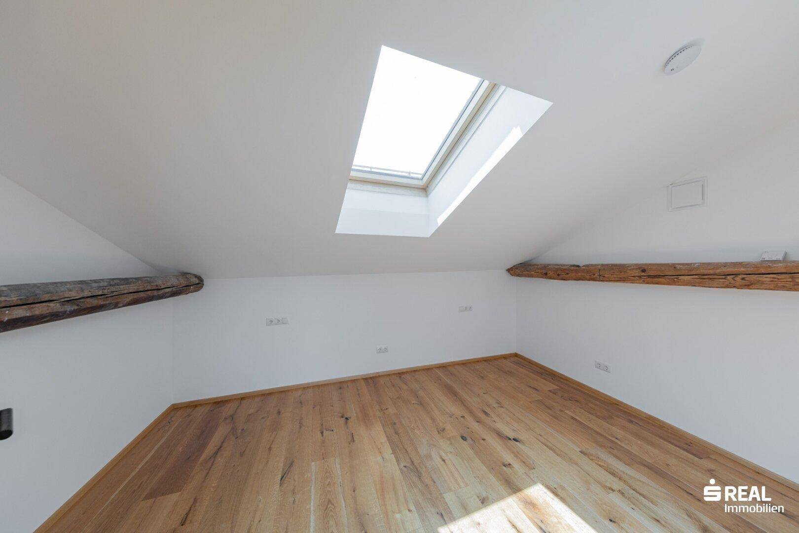 Dachgeschossraum mit Holzbalken, Parkettboden und einem großen Dachfenster für viel Licht.