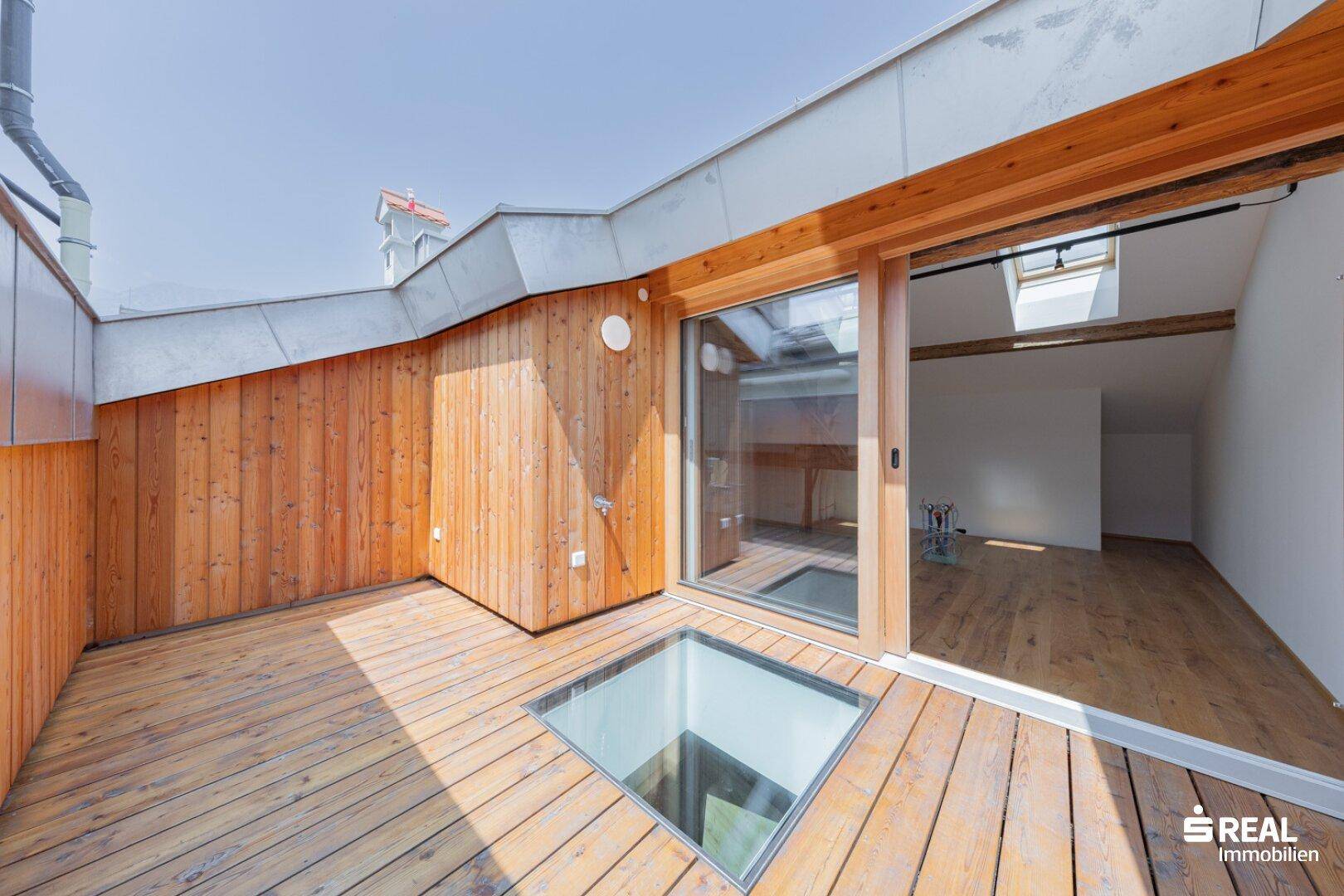 Sonnige Dachterrasse mit Holzbelag und Zugang zu einem hellen Innenraum.