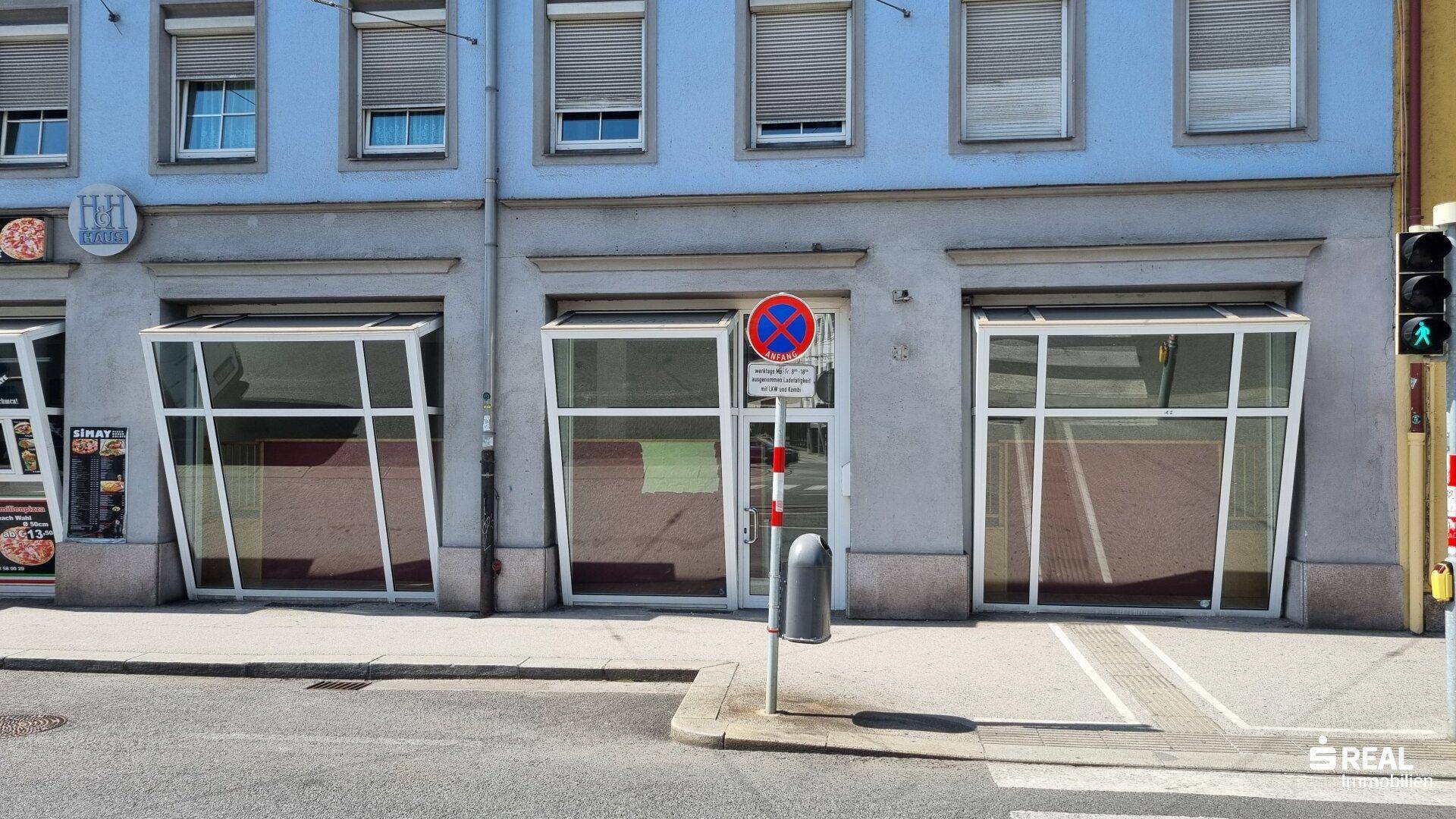 Nahaufnahme der Ladenfronten eines Gebäudes mit großen Glasflächen und Rollläden in der Stadt.