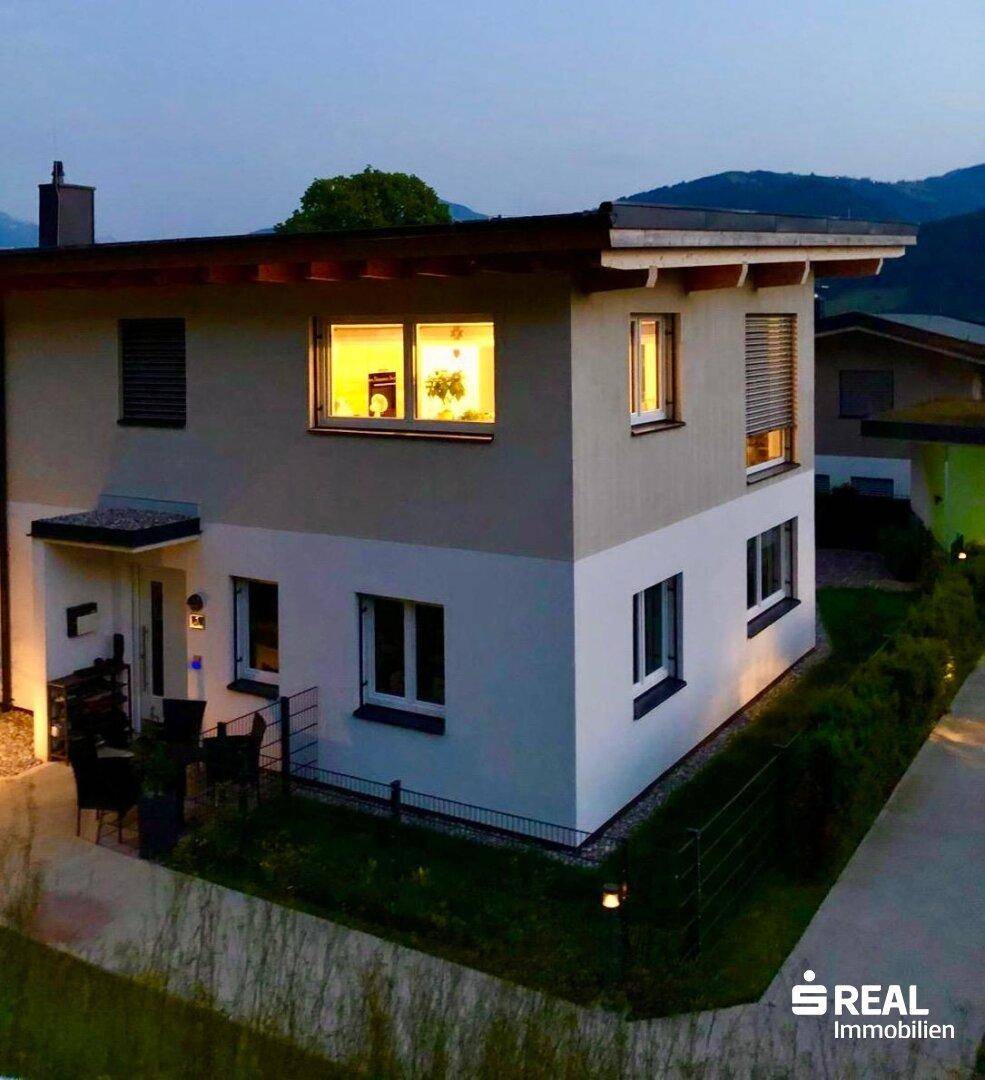 Modernes Einfamilienhaus bei Sonnenuntergang mit beleuchtetem Eingangsbereich und gepflegter Außenanlage.