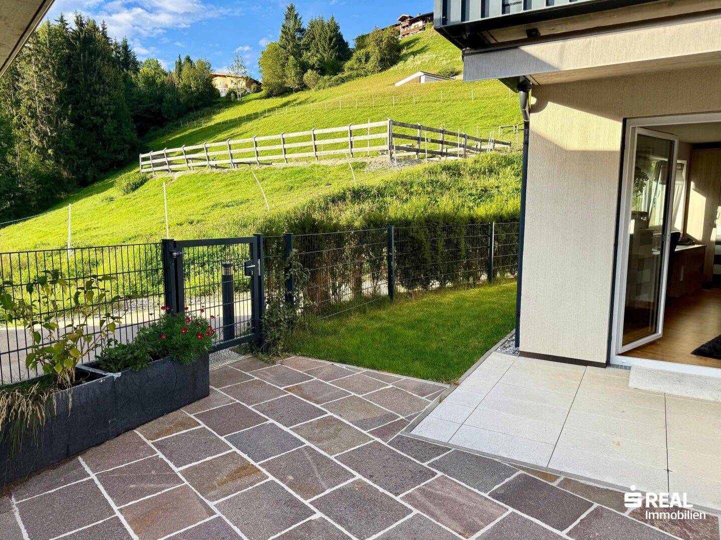 Gepflasterte Terrasse mit Zugang zum Garten und Blick auf die grüne Hanglage.