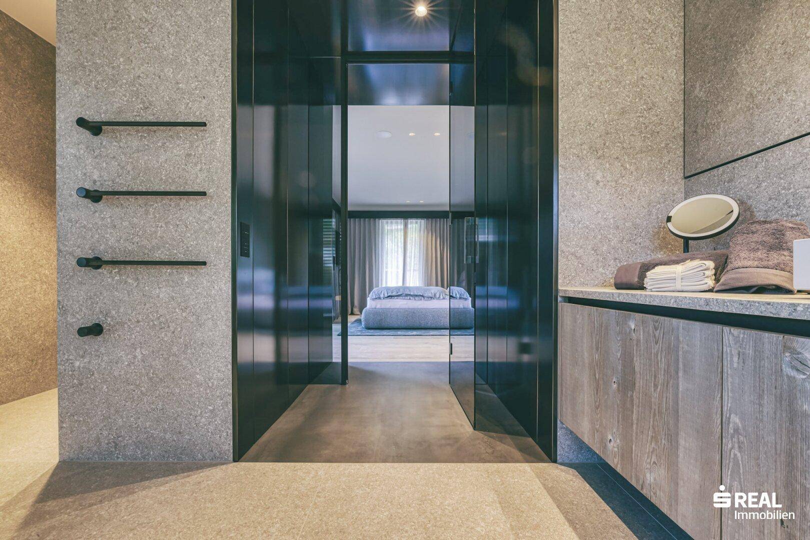 Modernes Badezimmer mit Steinfliesen und Blick durch Glastüren ins Schlafzimmer.