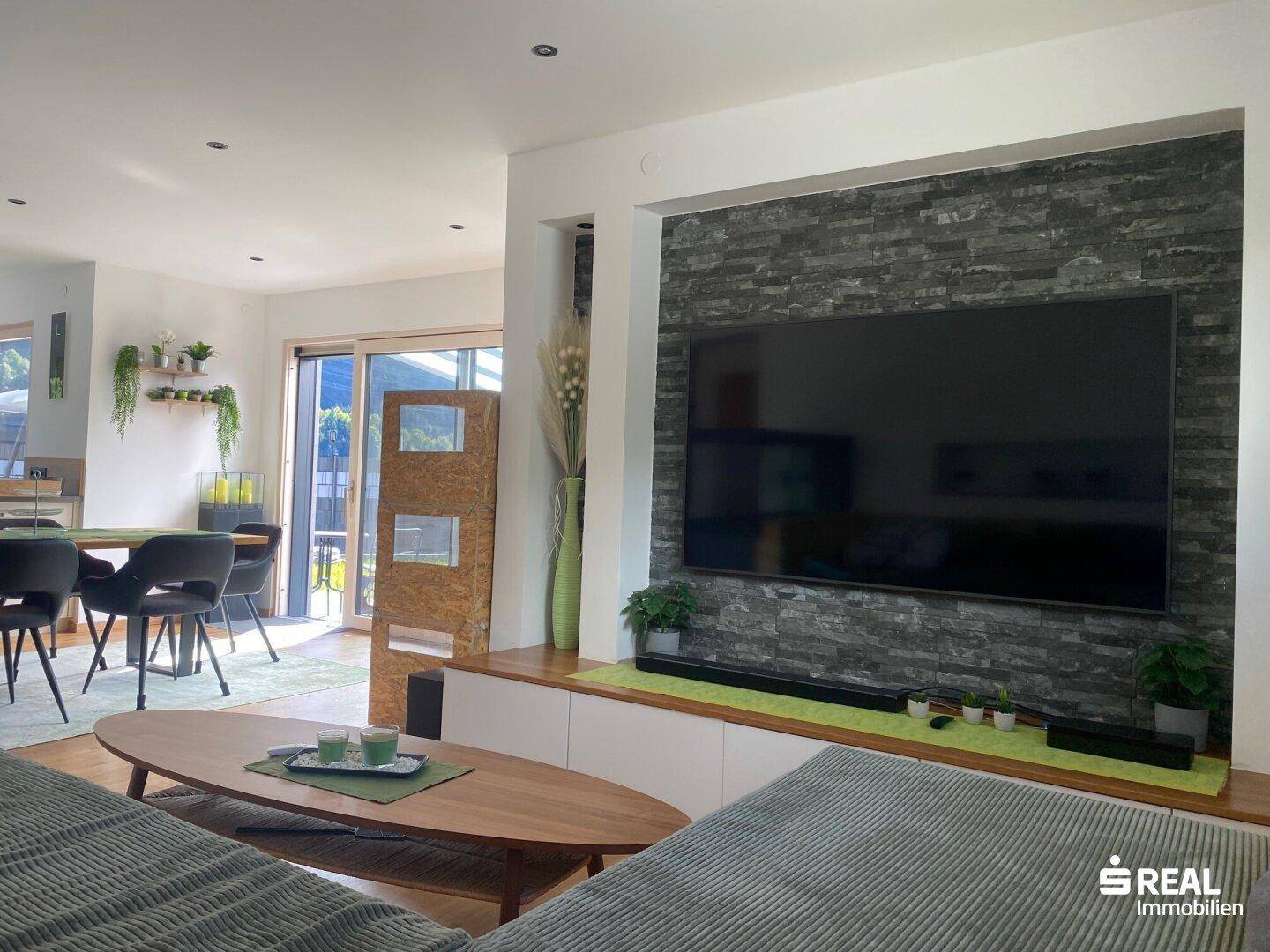 Geräumiger Wohnbereich mit großem Fernseher, Steinwand und Holzboden, offen zum Essbereich.