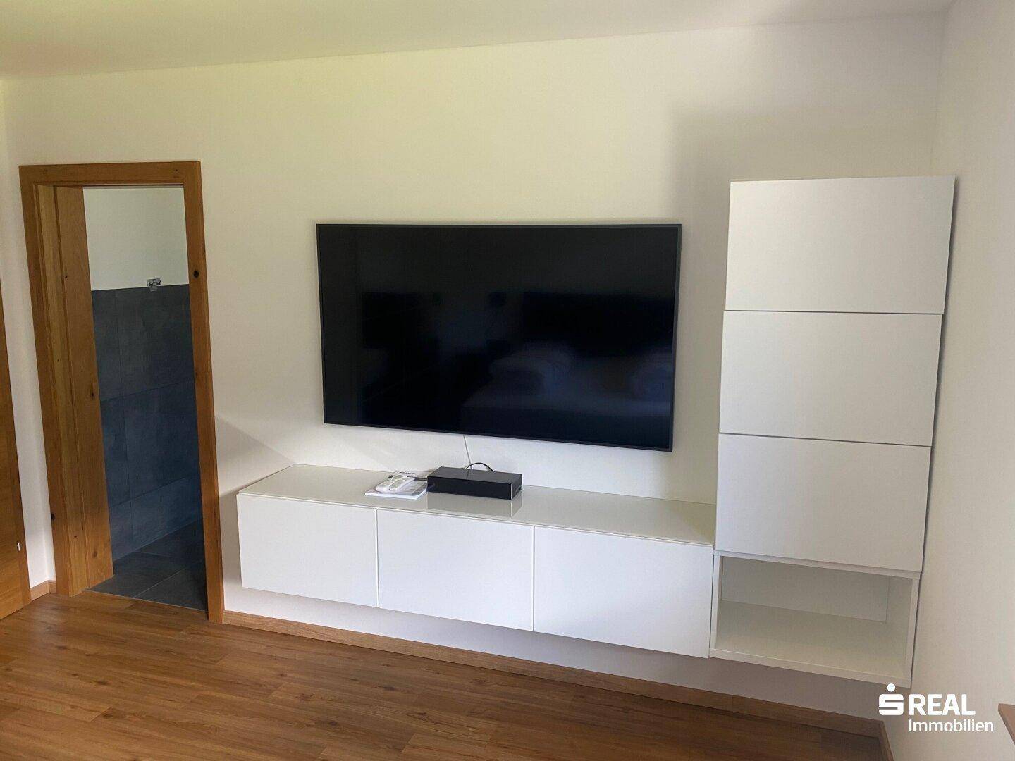 Wohnbereich mit Wandfernseher und weißen Schränken, Holzboden und schlichter Einrichtung.