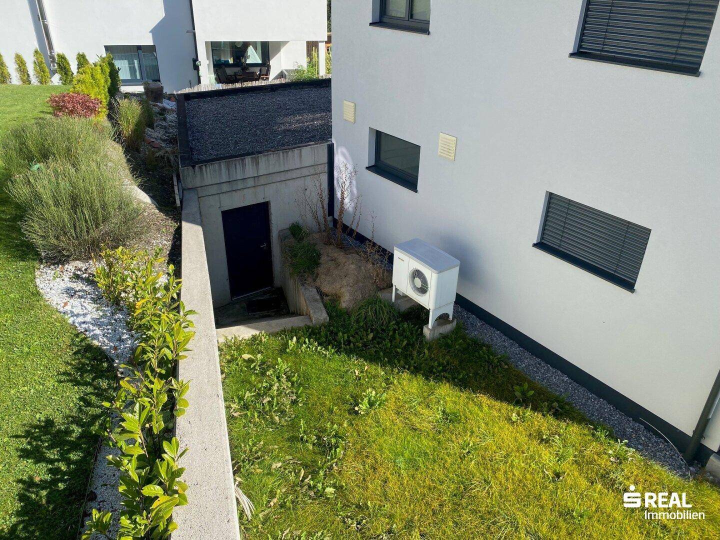 Seitlicher Gartenbereich mit Rasenfläche, Hecke, Außenschuppen und Wärmepumpe.