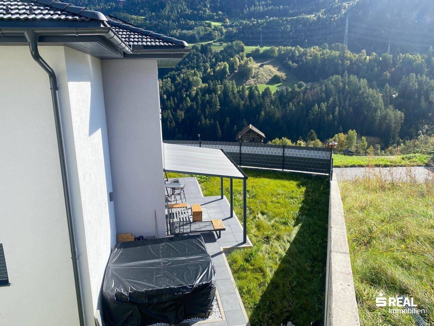 Überdachte Terrasse mit Sitzgelegenheiten und Blick auf die grüne Umgebung und Berge.