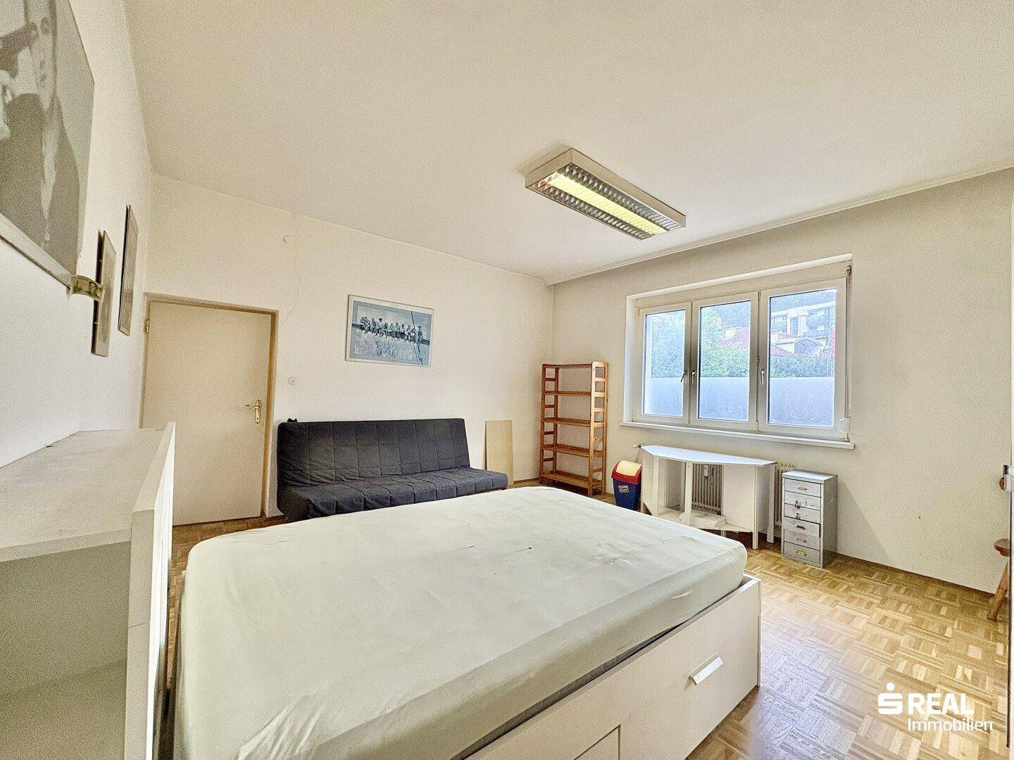 Schlafzimmer mit Parkettboden, großem Fenster und einfacher Einrichtung, bietet viel Licht.