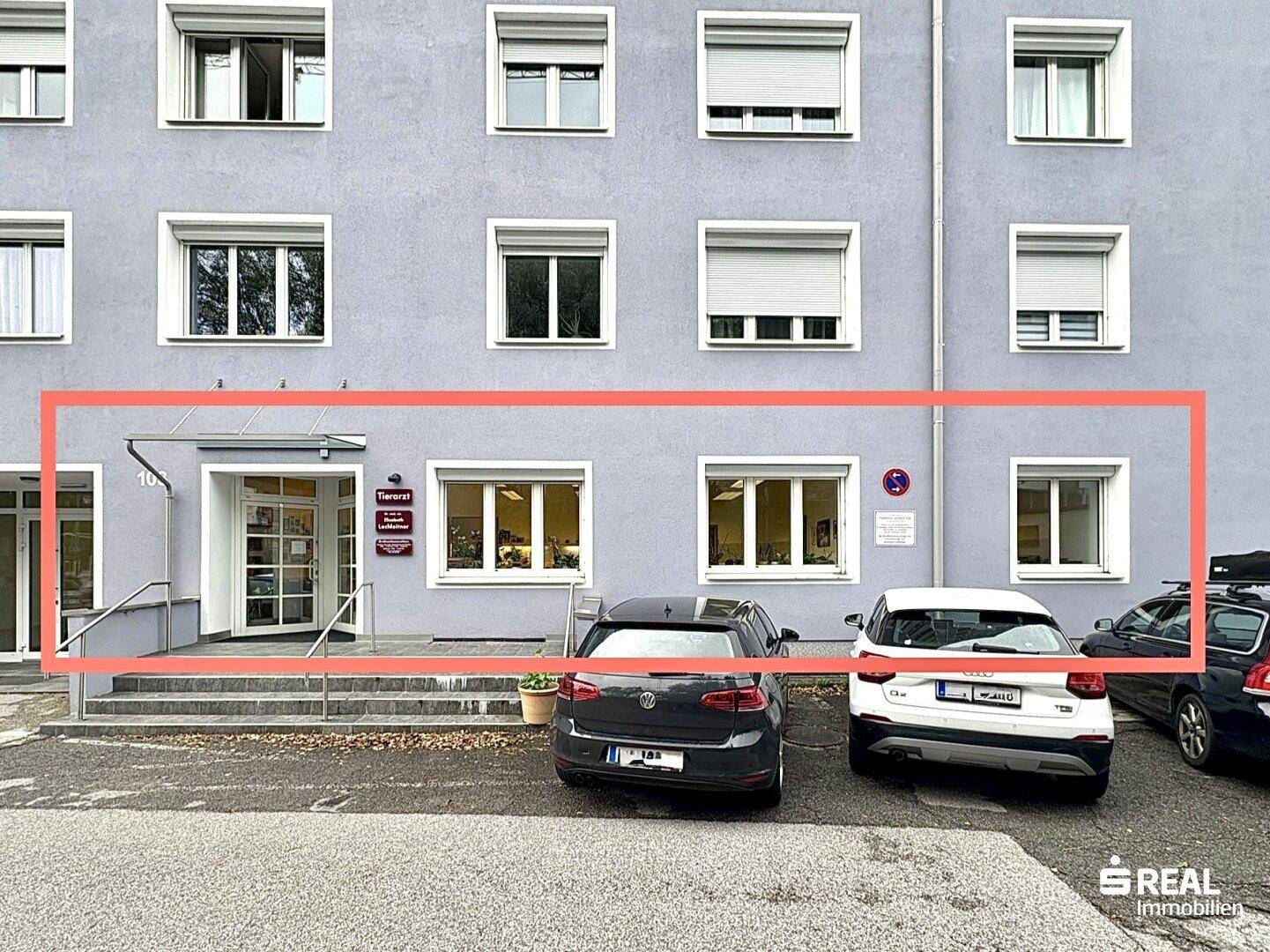 Graue Gebäudefassade mit mehreren Fenstern und Eingangsbereich, umrandet von parkenden Autos.