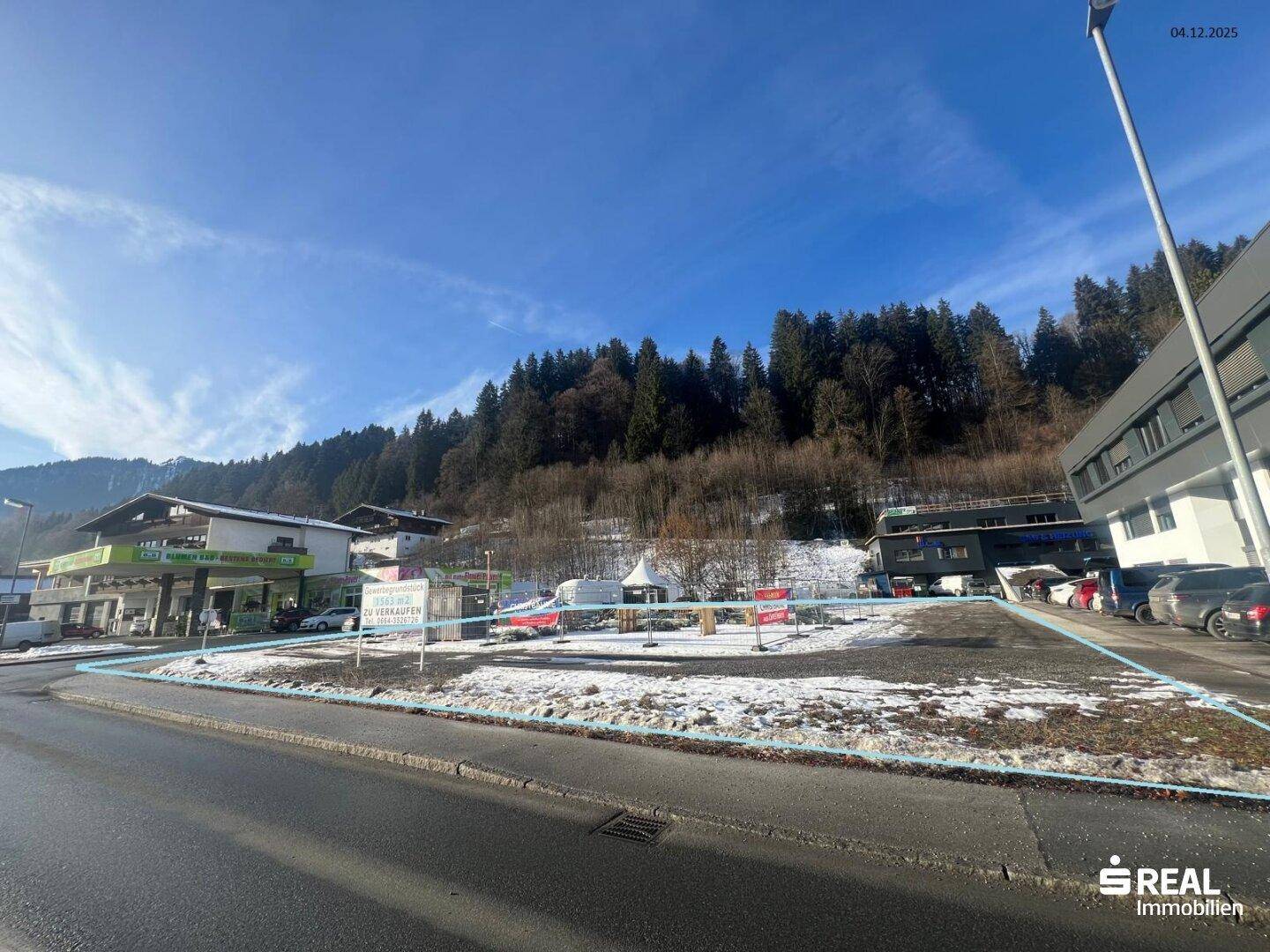 Blick von der Straße auf das verschneite Grundstück mit blauer Markierung und bewaldeten Hängen im Hintergrund.
