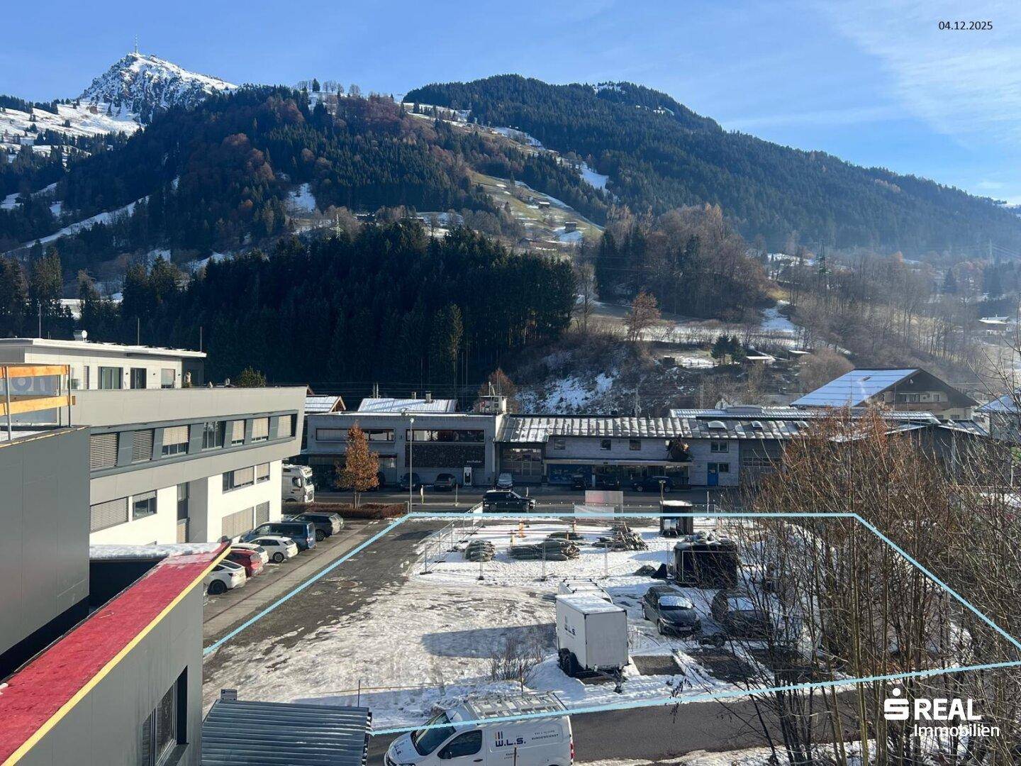 Winterliche Ansicht eines Grundstücks mit blauer Markierung, umgeben von verschneiten Bergen und Gebäuden.
