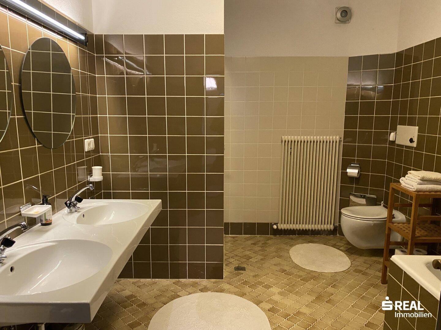 Geräumiges Badezimmer mit braunen und beigen Fliesen, Doppelwaschbecken und Heizkörper.