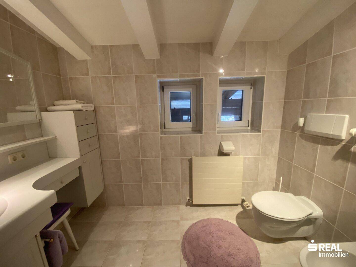 Helles Badezimmer mit beigefarbenen Fliesen, Waschtisch und Toilette, gut beleuchtet durch kleine Fenster.