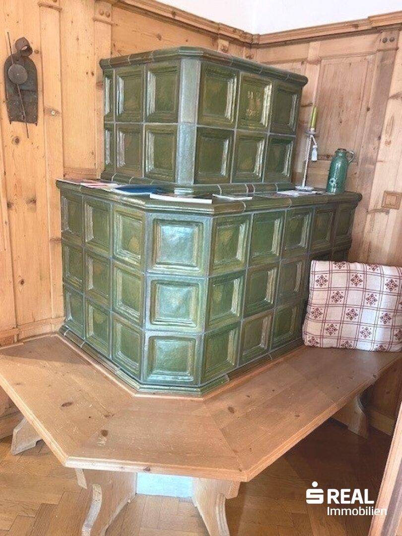 Traditioneller Raum mit grünem Kachelofen und Holztisch, umgeben von Holztäfelung.
