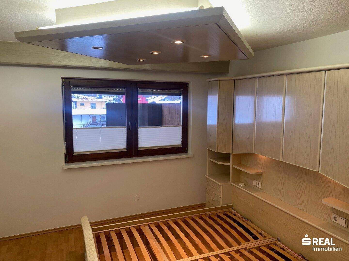 Schlafzimmer mit hellem Holzbettrahmen und praktischen Einbauschränken unter einem Fenster.