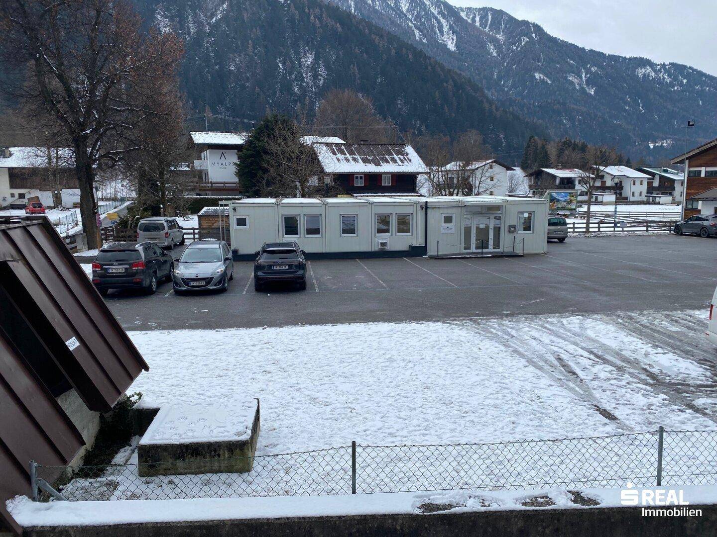 Außenparkplatz mit mehreren Fahrzeugen und temporären Gebäuden vor einer Bergkulisse.