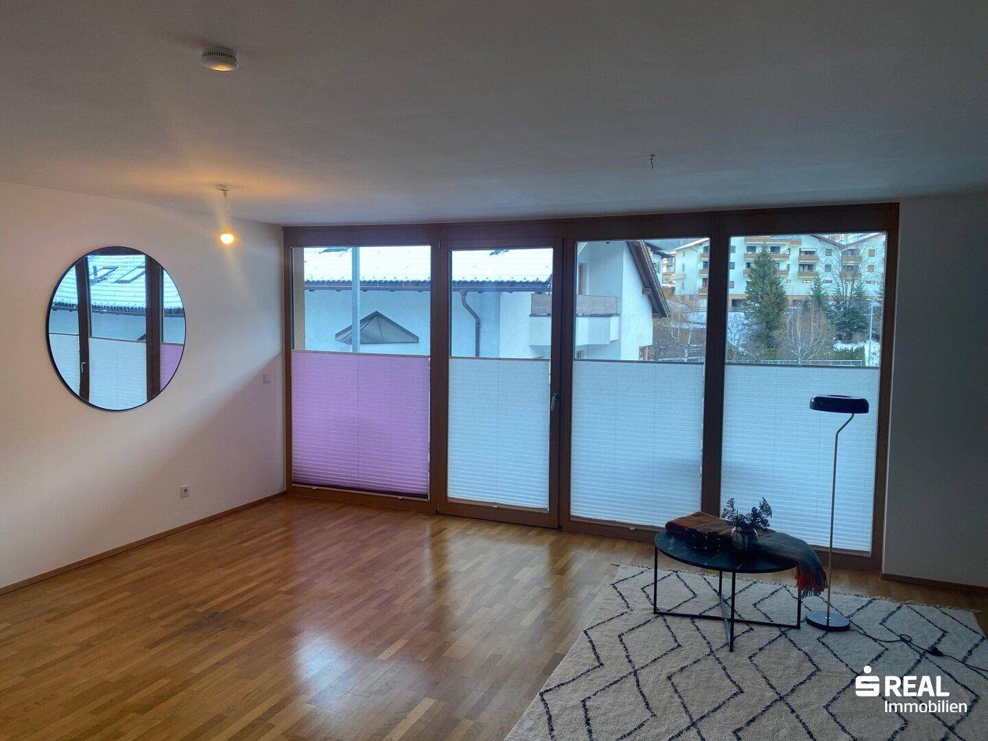 Geräumiges Wohnzimmer mit Holzboden und einer Fensterfront, die viel Licht hereinlässt.