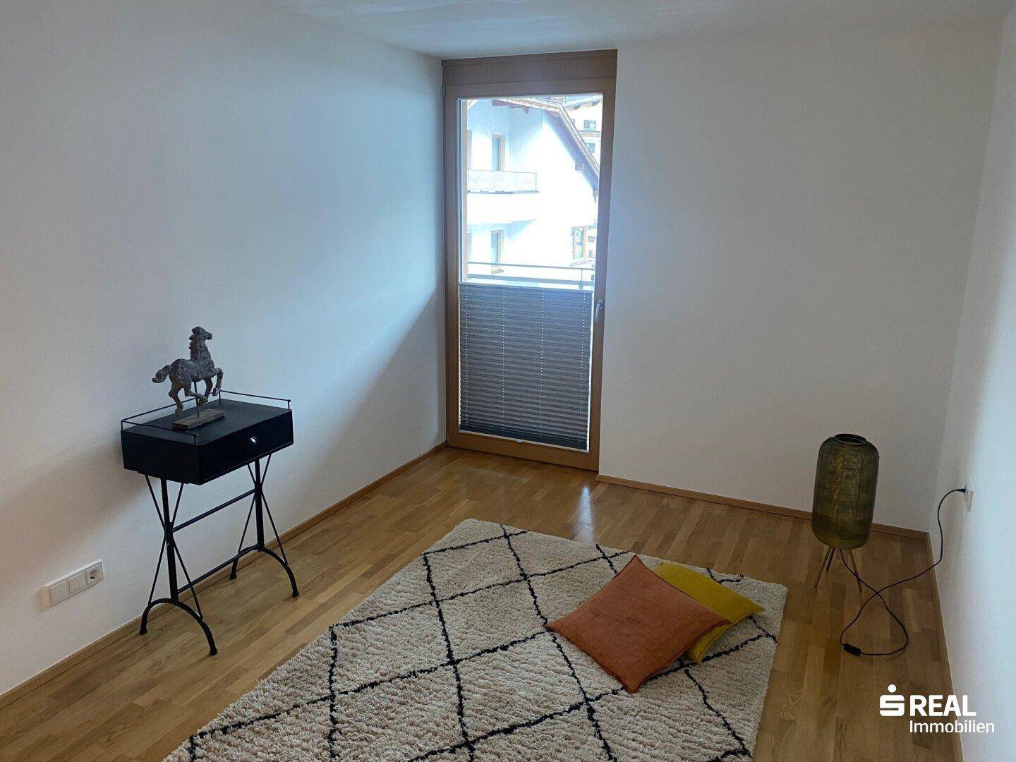 Leeres Zimmer mit Holzboden, großem Fenster und einfacher Einrichtung.