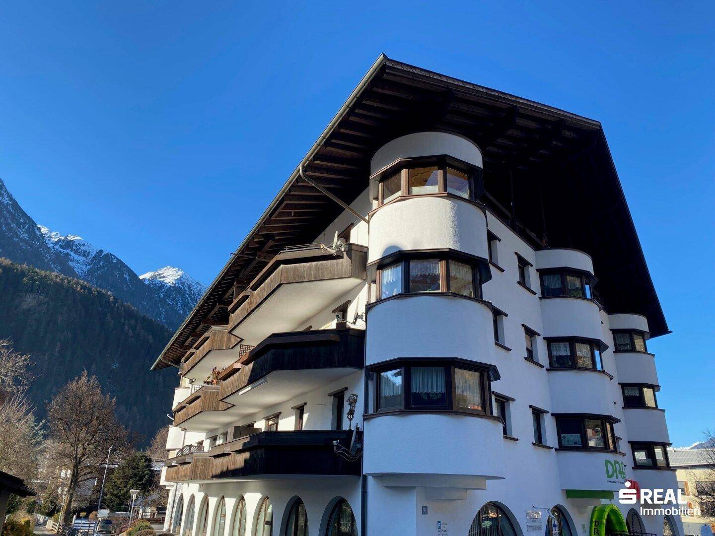 Mehrstöckiges Gebäude mit traditioneller Architektur, Erkerfenstern und Blick auf die umliegenden Berge.