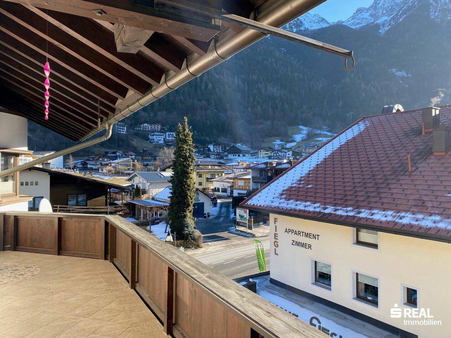 Balkon mit Blick auf die Dorfstraße und die umliegende Berglandschaft unter blauem Himmel.