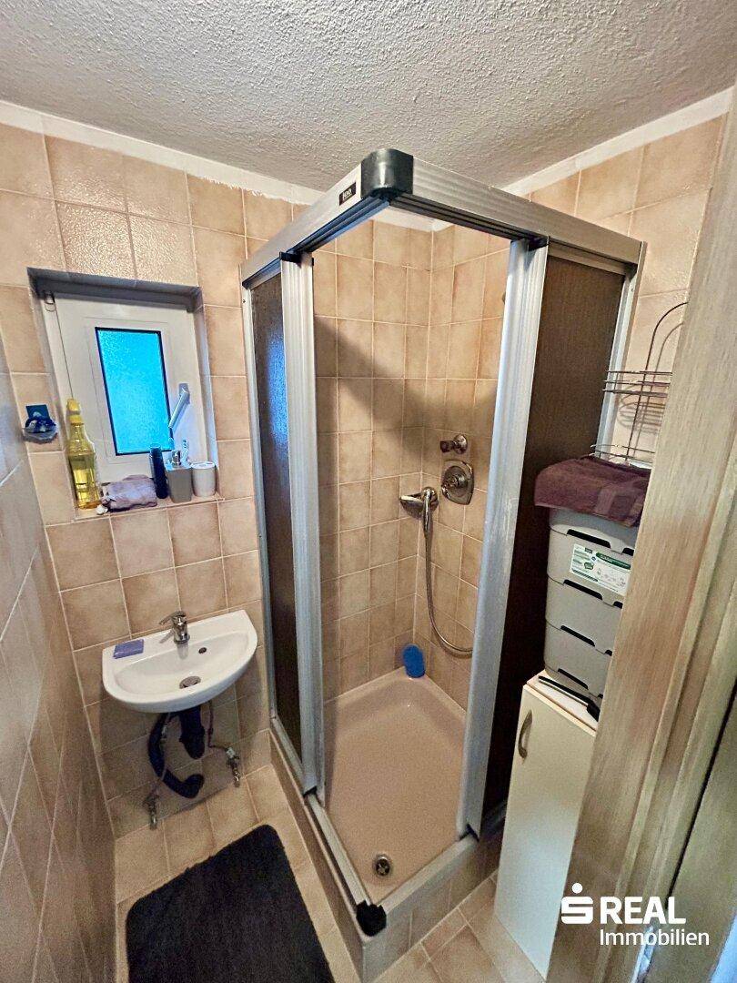 Badezimmer mit Duschkabine, kleinem Waschbecken und hellbraunen Fliesen an den Wänden.