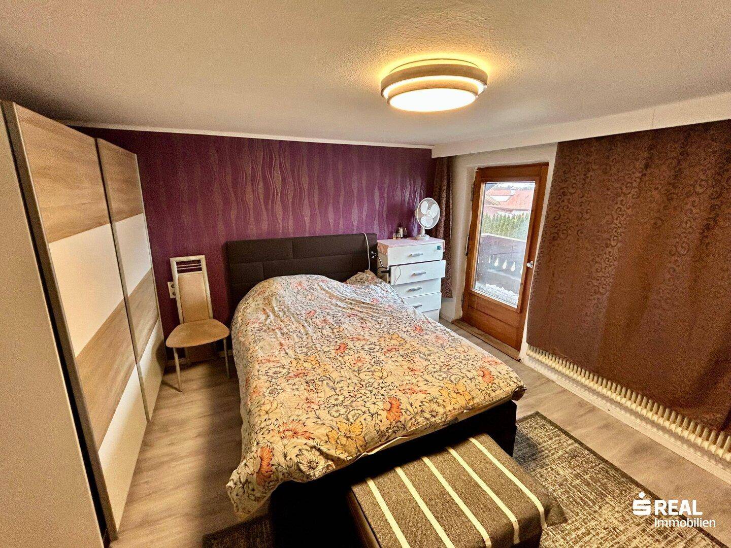 Schlafzimmer mit Doppelbett, violetter Akzentwand und Zugang zu einem Außenbereich.