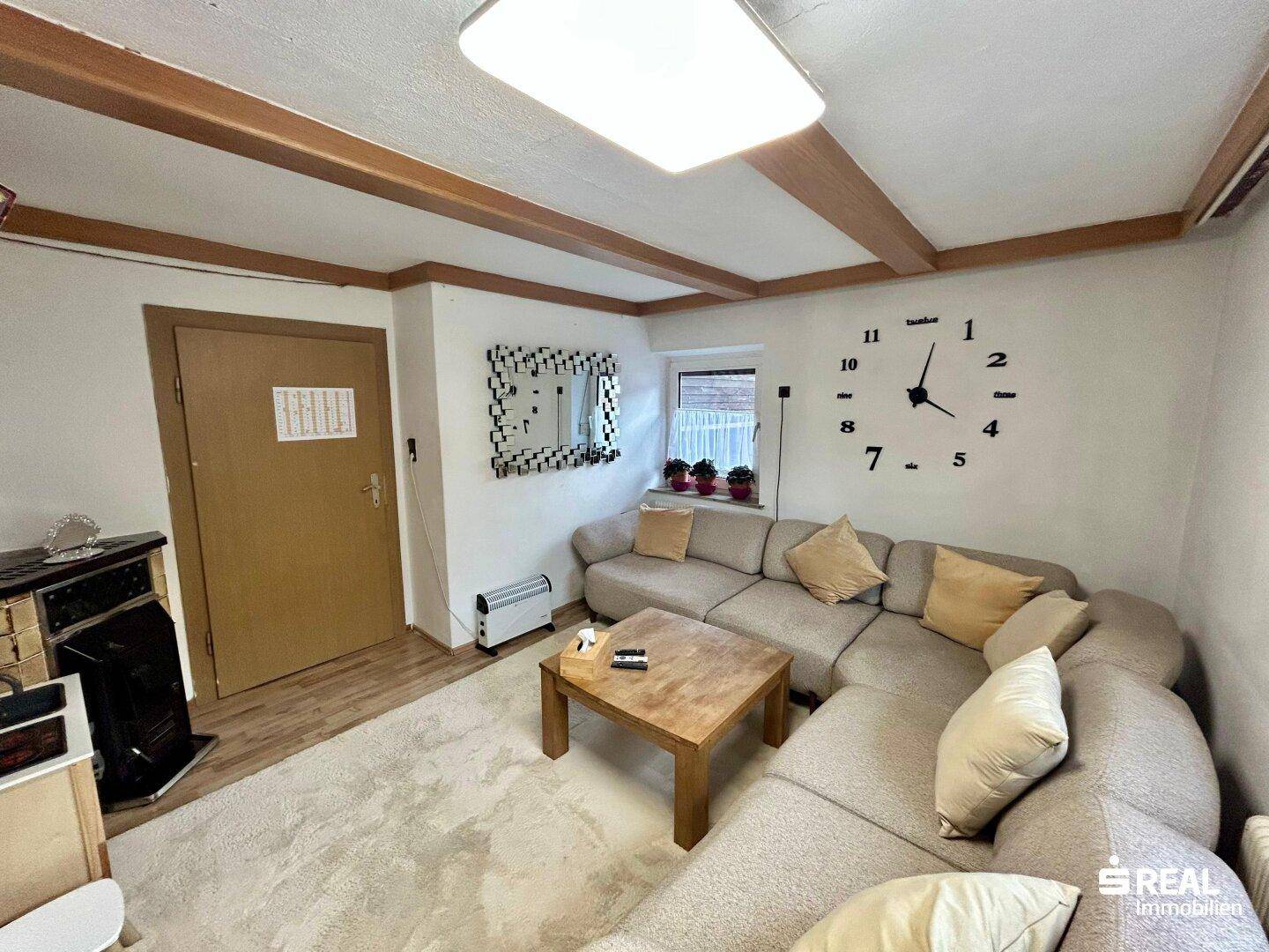 Gemütliches Wohnzimmer mit großem Ecksofa, Holztisch und dekorativer Wanduhr unter Holzbalken.