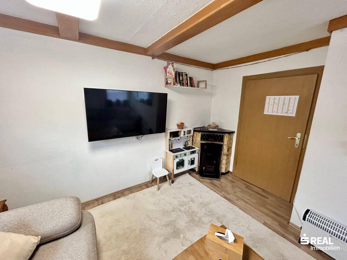 Wohnzimmer mit wandmontiertem Fernseher und einem kleinen Spielbereich, ergänzt durch Holzbalken.