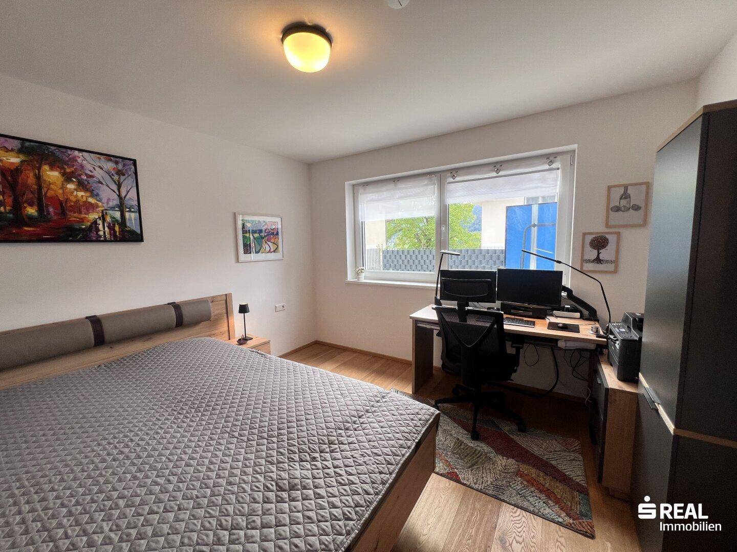 Schlafzimmer mit Holzboden, großem Bett und Arbeitsbereich am Fenster.
