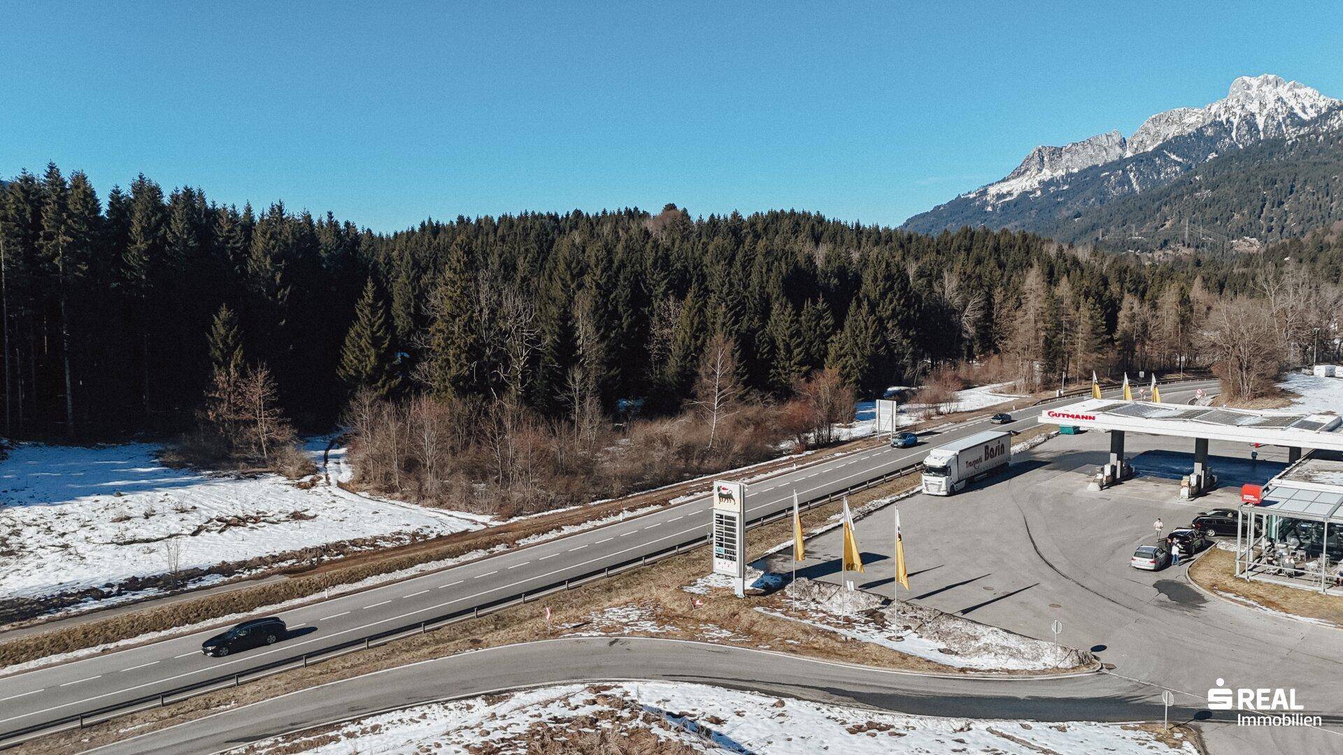 Weitläufige Landschaft mit einer Straße, Tankstelle, Wald und schneebedeckten Bergen im Hintergrund.