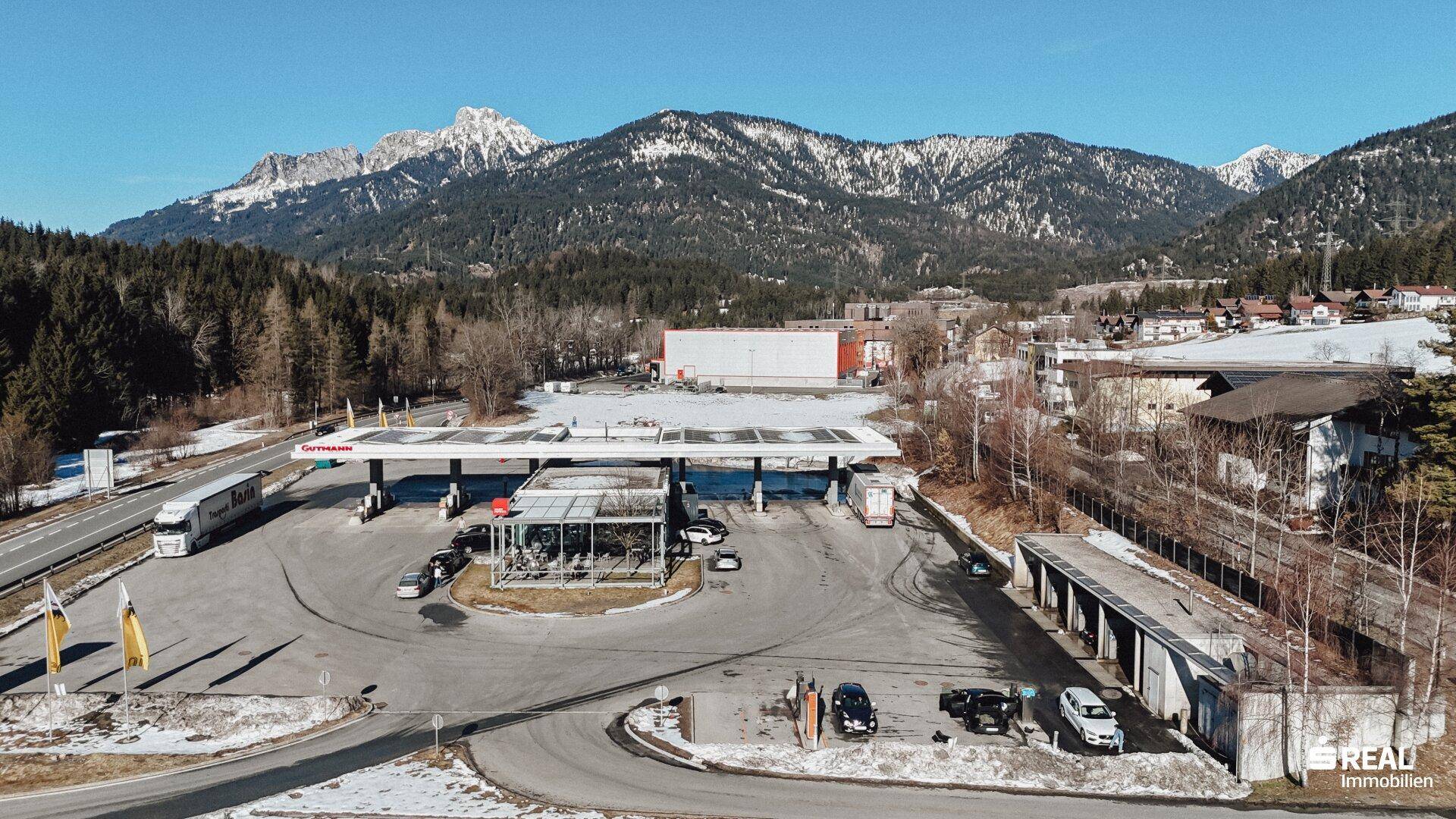 Luftaufnahme einer Tankstelle an einer Straße mit schneebedeckten Bergen und Wald im Hintergrund.
