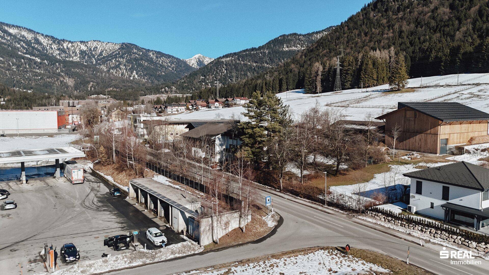 Blick auf eine Tankstelle und angrenzende Wohnhäuser in einer bergigen, teilweise verschneiten Landschaft.