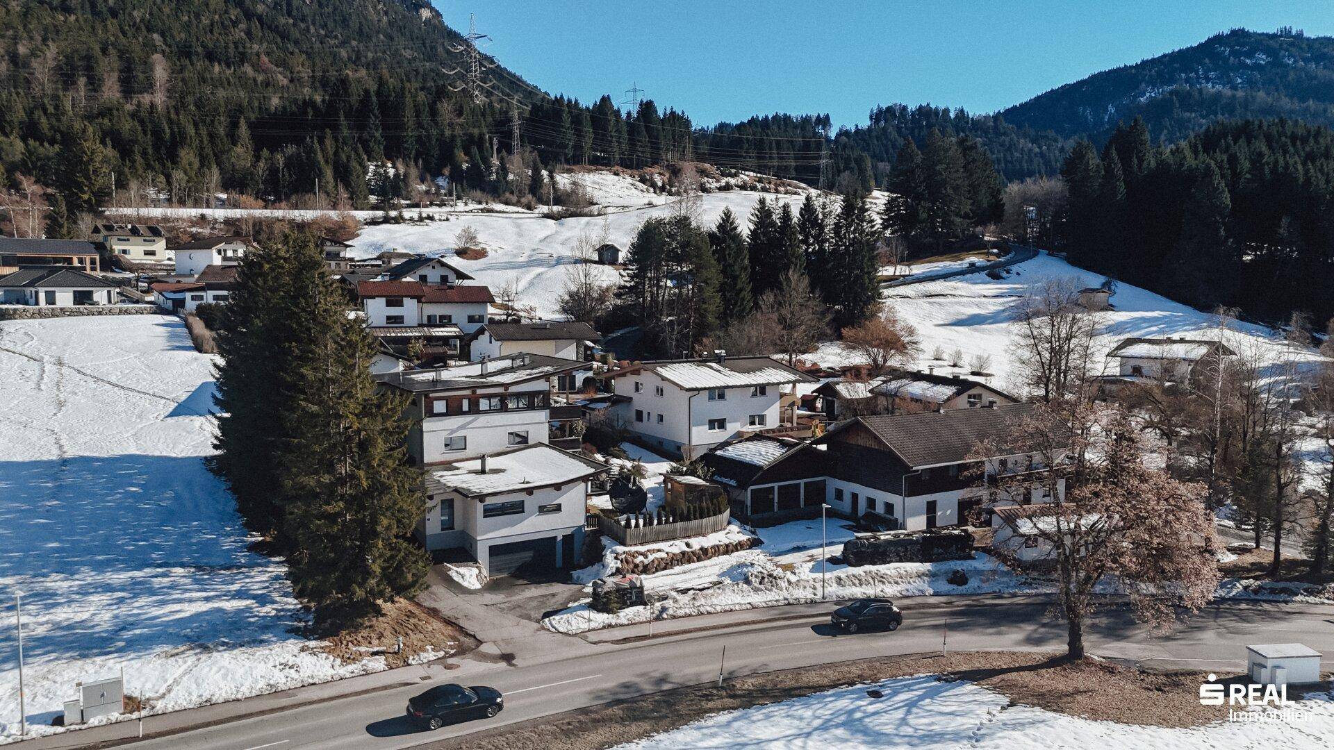 Wohnsiedlung mit mehreren Häusern entlang einer Straße in einer winterlichen Umgebung mit Bergen.