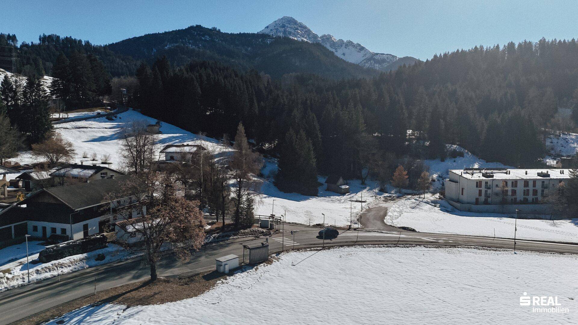 Panoramablick auf eine ländliche Gegend mit Häusern, Wald und schneebedeckten Bergen im Sonnenschein.