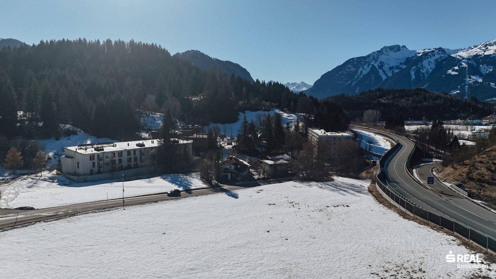 Weitläufige Landschaft mit einer Straße, Gebäuden, Wald und majestätischen Bergen im Hintergrund.