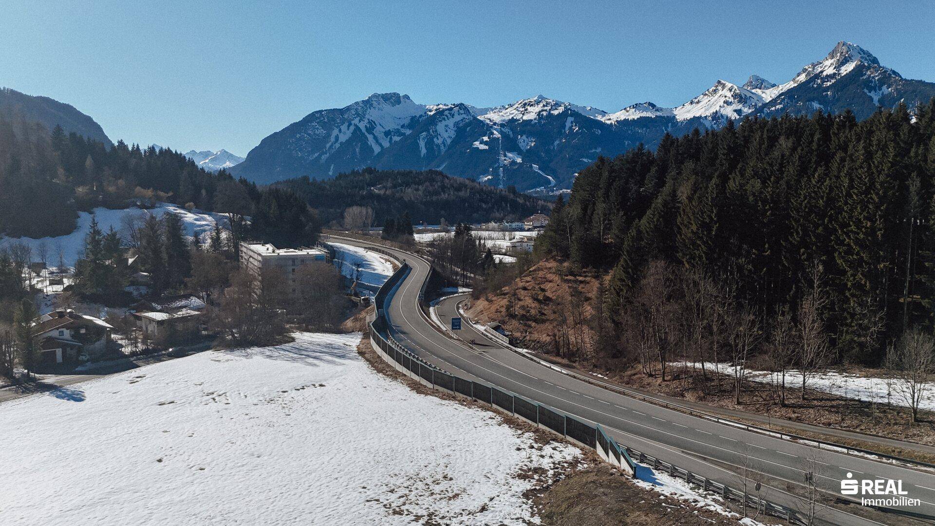 Autobahnabschnitt schlängelt sich durch eine bewaldete Landschaft mit schneebedeckten Bergen unter blauem Himmel.