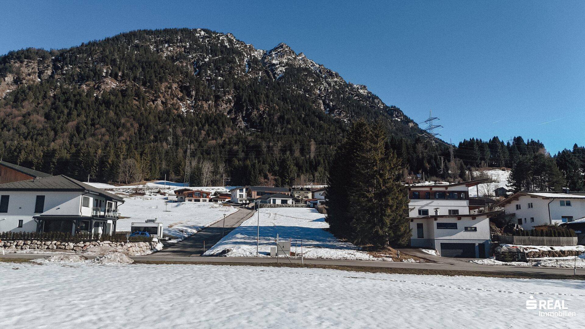 Wohngebiet mit modernen Häusern und einem verschneiten Feld vor einer bewaldeten Bergkulisse.