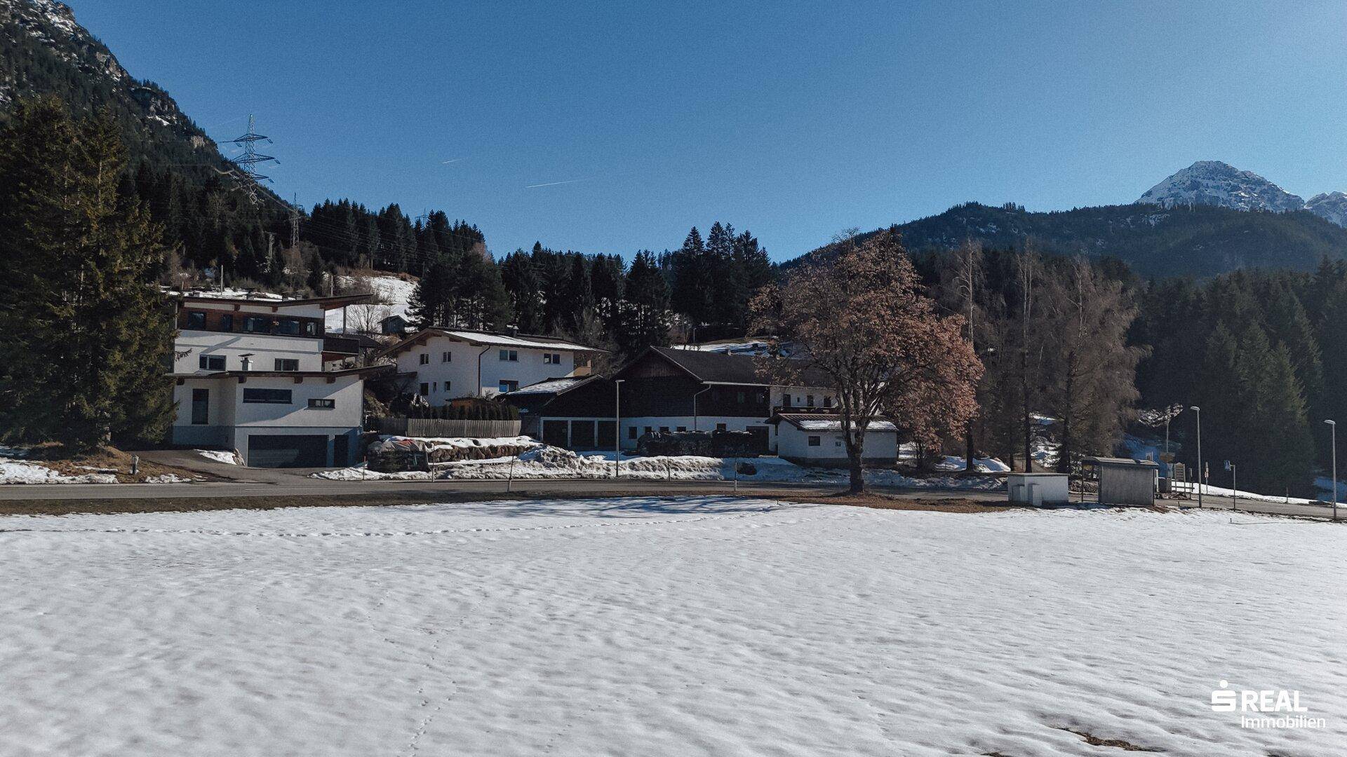 Ländliche Wohngegend mit Häusern und einem weiten, verschneiten Feld vor den Bergen.