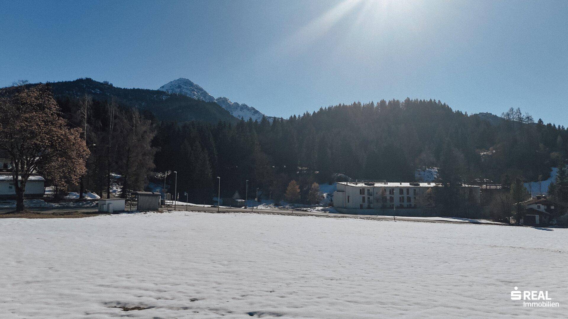 Weitläufiges, verschneites Feld mit Gebäuden, Wald und schneebedeckten Bergen in der Ferne.
