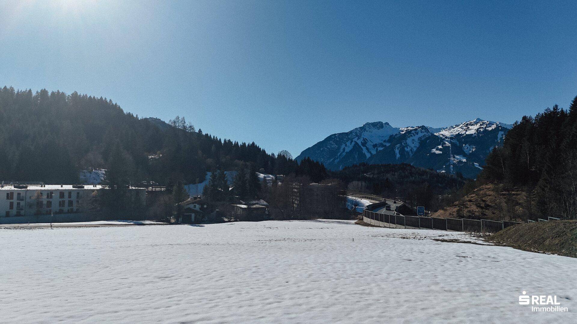 Panoramablick über ein verschneites Feld mit Gebäuden, Wald und einer imposanten Bergkette.