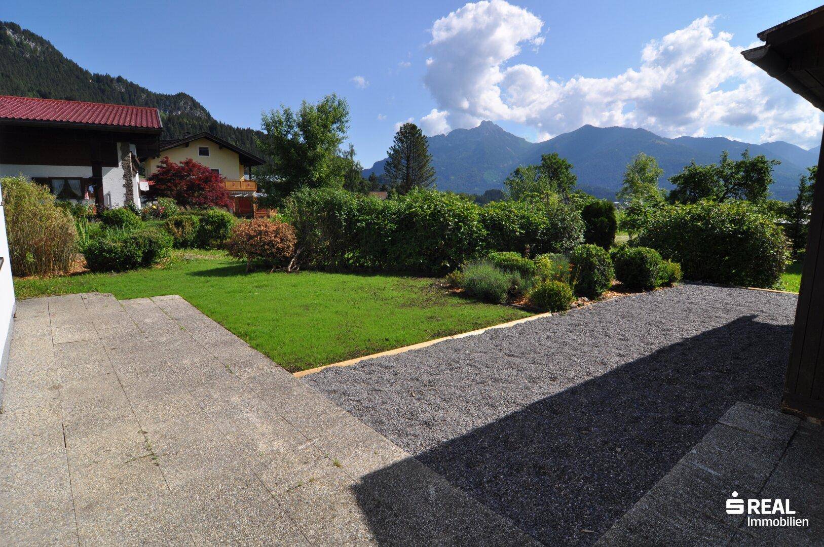 Gepflegter Gartenbereich mit Rasenfläche, gepflasterter Terrasse und Hecken im Hintergrund.