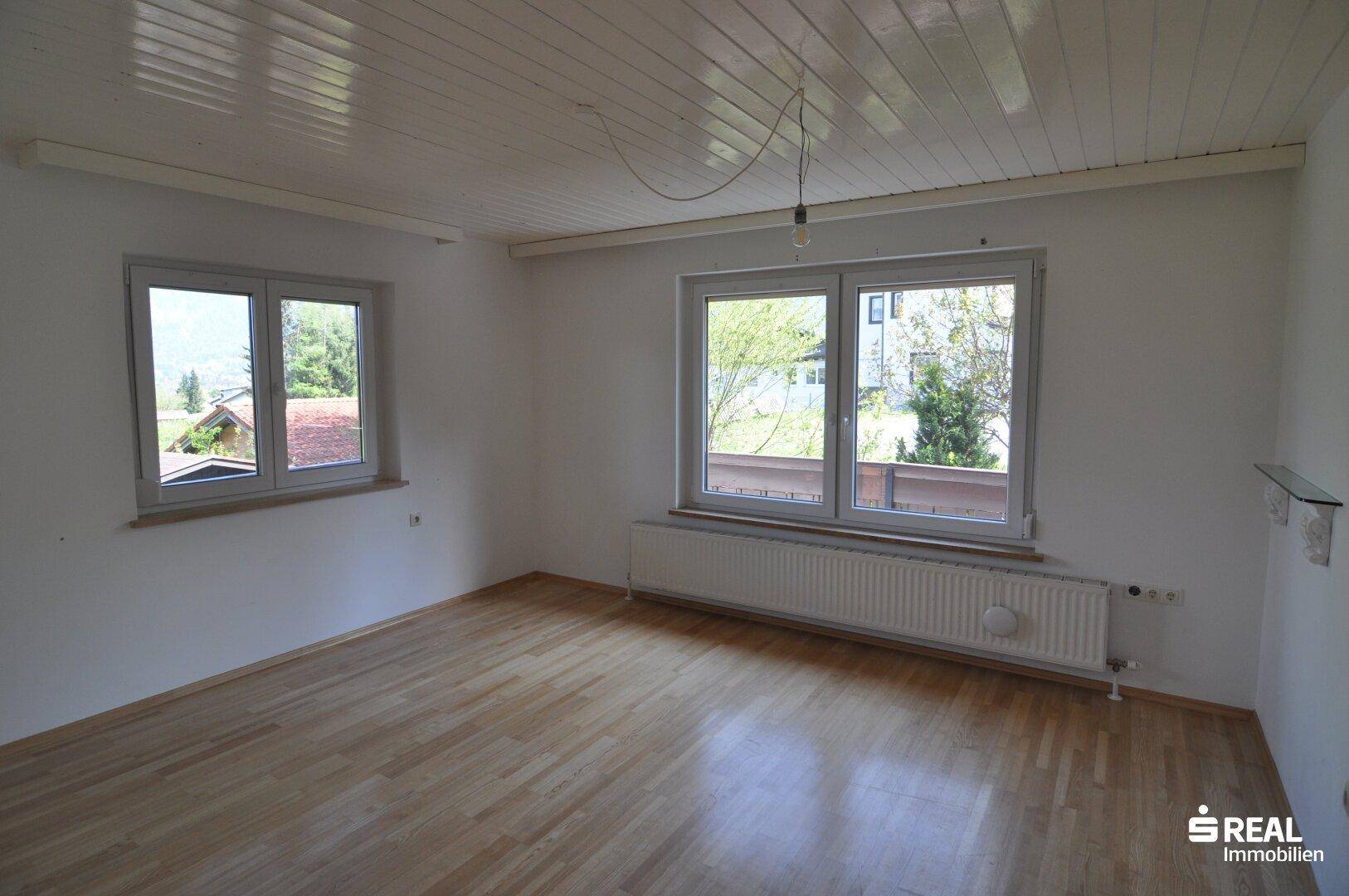 Geräumiger Raum mit Holzparkettboden, großen Fenstern und Blick ins Grüne.
